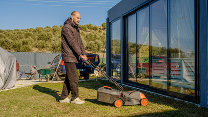 A middle-aged man mowing the grass in his garden with a lawnmower on a winter day