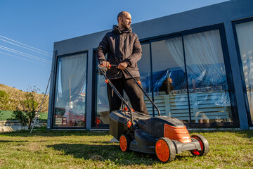 A middle-aged man mowing the grass in his garden with a lawnmower on a winter day