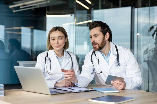 Medical professionals working together at an office desk, discussing information on a laptop, a male doctor pointing at the screen while holding a digital tablet - Powered by Adobe