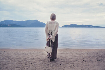 Woman in a white crochet sweater standing on a sandy lakeshore, looking out at calm water and mountains.