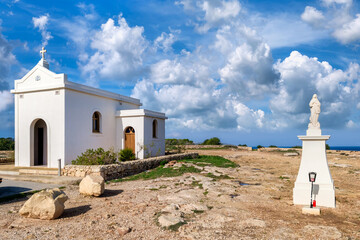 Chapel of the Immaculate Conception with a statue of a saint in Mellieha, Malta, on a rocky plateau overlooking the sea under a clear blue sky.