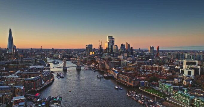 Aerial panorama of London skyline at sunset, with Tower Bridge crossing the River Thames and city landmarks bathed in warm autumn light during dusk. Drone flight over modern cityscape perspective