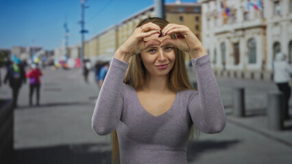 Fototapeta premium Woman making heart gesture on city street with long blonde hair wearing gray shirt smiling outdoors in urban environment with people and buildings in background showing love and happiness.