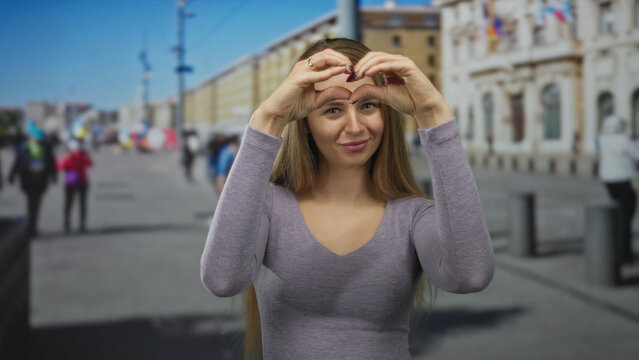 Woman making heart gesture on city street with long blonde hair wearing gray shirt smiling outdoors in urban environment with people and buildings in background showing love and happiness.