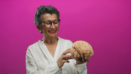 Senior woman with grey hair examines brain model against bright pink background, showcasing...