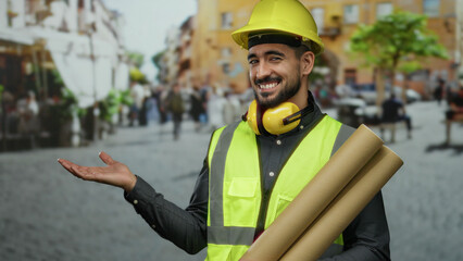 Young man in a hard hat and vest stands confidently on an urban street, extending one hand while...