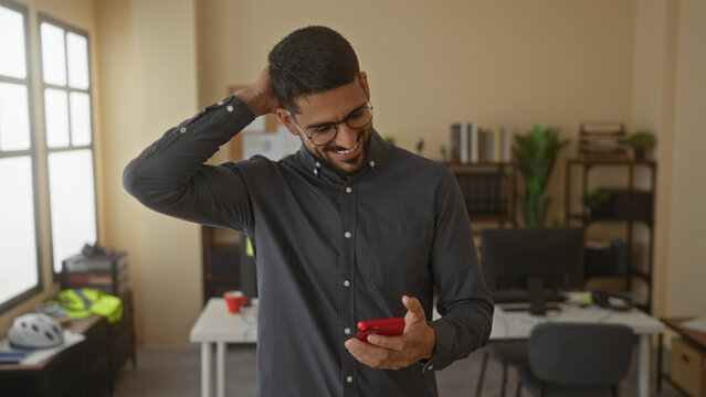 Young man with glasses smiles while using smartphone in modern office featuring bright decor and shelves, creating a professional and relaxed work atmosphere.