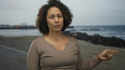 Woman with curly hair finger pointing at sandy beach shoreline with ocean horizon visible; determination.