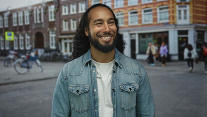 Man with long curly hair and beard smiling and looking up on a busy city street in front of a...