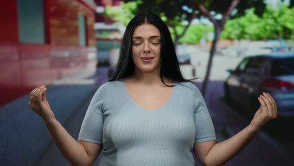 Woman meditating outdoors in a city street poses peacefully wearing casual attire with hands together, embracing tranquility surrounded by urban trees, cars, and buildings.