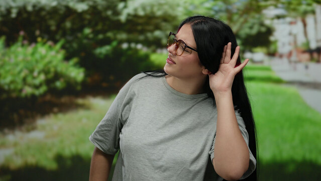 Woman standing in a park wearing glasses and a grey shirt with a hand gesture under the leafy green trees.