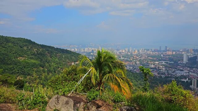 Panoramic view of Penang city and George Town from Penang Hill viewpoint. Urban landscape with high-rise buildings, mountains and tropical greenery under blue sky with clouds. Scenic overlook of islan