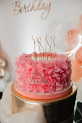 Beautiful pink ruffled birthday cake with golden candles and Happy Birthday topper against a background of balloons.