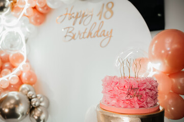 Beautiful pink ruffled birthday cake with golden candles and Happy Birthday topper against a background of balloons.