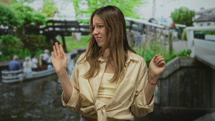 Woman raises both hands palms out by canal on street in city, shrugging with a slight grimace; bemusement hesitation.