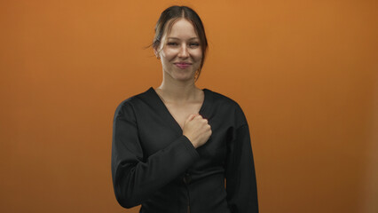Fototapeta premium Young hispanic woman with clenched fist punching forward, smiling, wearing black top in orange studio; determination empowerment.