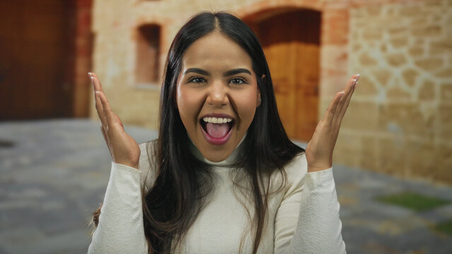 Young latin woman playfully covering face then smiling in old town setting showing joy and surprise outdoors with stone buildings as background