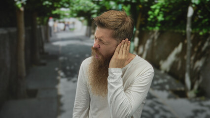 Caucasian man with long beard holds ear with hand in pain gesture on sunlit city street lined with...