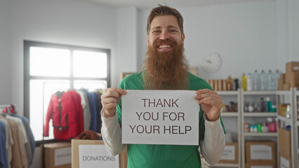 Bearded smiling man holding sign with both hands at donation center building with shelves of boxes and racks; gratitude.