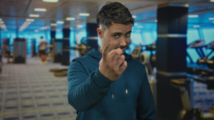 Young man in a gym wearing a hoodie glancing at the camera with fitness equipment blurred in the background, suggesting an indoor sports environment.