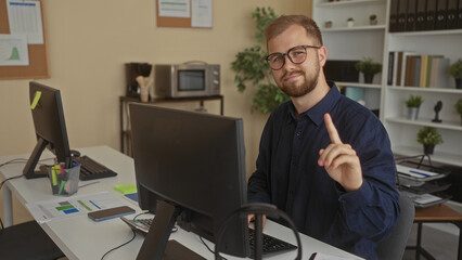 Young man typing at keyboard and pointing index finger while seated at desk with monitor, smartphone and documents in an office building; concentration productivity.