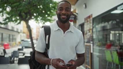 Man smiling and using smartphone while walking down a city street with backpack and bracelet visible; confidence connection ease.