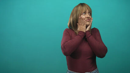 Woman with hands on cheeks and expressive shrug gestures in studio with teal backdrop, maroon top and jeans, six shot sequence shows shifting surprise and pondering; pensive reflection.