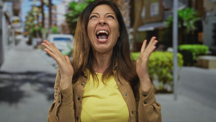 Hispanic middle age woman claps raised hands with open mouth on sunny city street amid blurred storefronts and potted shrubs; joy.