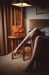 Elegant orange leather handbag on a side table next to a woman relaxing in an armchair under warm lamp light.