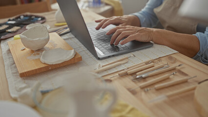 Middle aged caucasian woman types on laptop beside white clay bowl and wooden carving tools on...