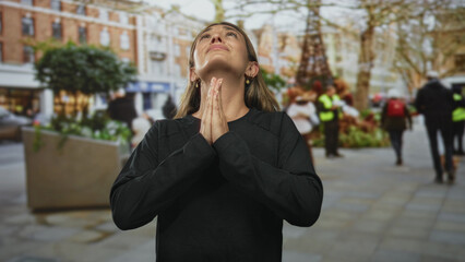 Woman with hands pressed together in prayer, head tilted up on a city street amid pedestrians; hope.