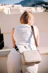 Woman in a white linen suit leaning on a balcony overlooking a Mediterranean coastal town with a beige bag.