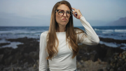 Woman points finger to temple while adjusting glasses on rocky seaside coast near waves, outdoors; puzzled thought.
