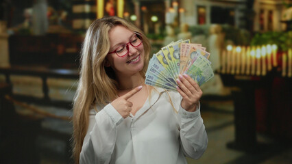 Woman smiling indoors at a church with romanian banknotes, wearing glasses and pointing happily at...
