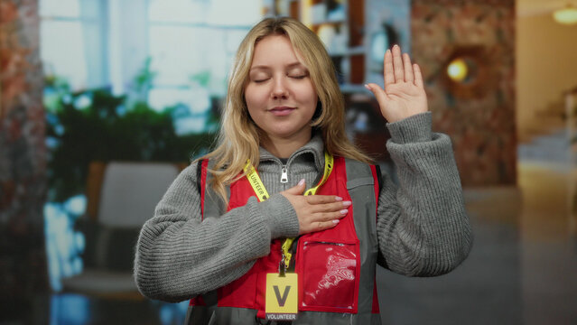 Woman volunteer in hotel gestures an oath with hand on heart wearing red vest indoors as she expresses commitment and dedication.