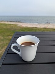 Close-up of a white mug filled with coffee standing on a wooden table in the garden overlooking the sea