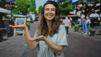 Fototapeta premium Young woman in light blue dress smiles while holding both open palms on a busy street; optimism hope success ambition.