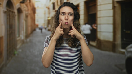 Fototapeta premium Woman pointing finger to cheeks against a sunlit old town street with curious expression; playful mischief.