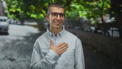 Man outdoors on a street with trees, wearing glasses and a striped shirt, smiling with hand on chest, creating a friendly urban atmosphere under green foliage.