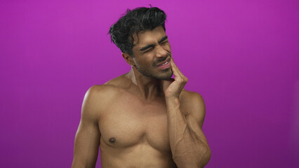 Fototapeta premium Shirtless young hispanic man with bare chest touches cheek with hand in purple studio; toothache pain.