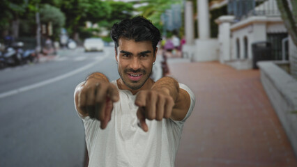Young hispanic man with smiling face points finger toward camera on city street during daytime; confidence.