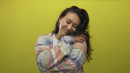 Woman hugging her shoulder in yellow studio wearing patterned sweater and hoop earrings, smiling with closed eyes and arms wrapped around herself; self love contentment.