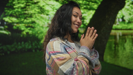 Fototapeta premium Woman with palms pressed together near chest in green park by a tree, smiling warmly and looking at viewer; joy gratitude.