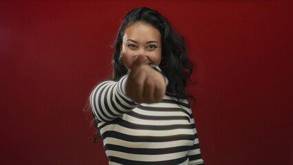Young hispanic woman smiling and making a finger heart gesture with fingers in studio against red wall; playful affection.