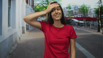 Young hispanic woman in red shirt with hand on forehead on a city street, palm to brow gesture; embarrassment.
