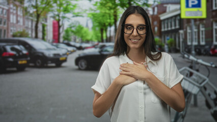 Young hispanic woman hands on chest smiling on a street with parked cars and a bicycle rack;...