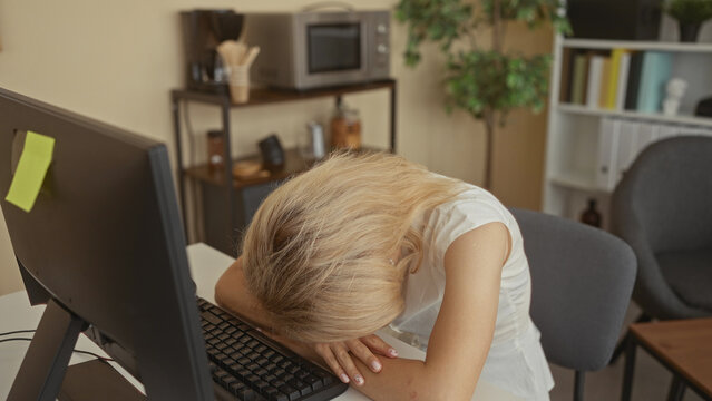 Young blonde woman rests forehead on white desk in modern office building at computer keyboard; tiredness.