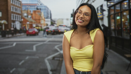 Woman smiling and looking up, showing cleavage in a yellow top on a street with storefronts and cars; joy confidence.
