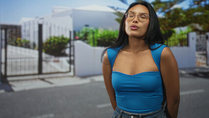 Woman with exposed chest and pursed lips wearing glasses standing on a street by a gated house; quiet contemplation.