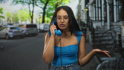 Young brunette woman holds vintage blue telephone handset to her ear on a city street and raises...